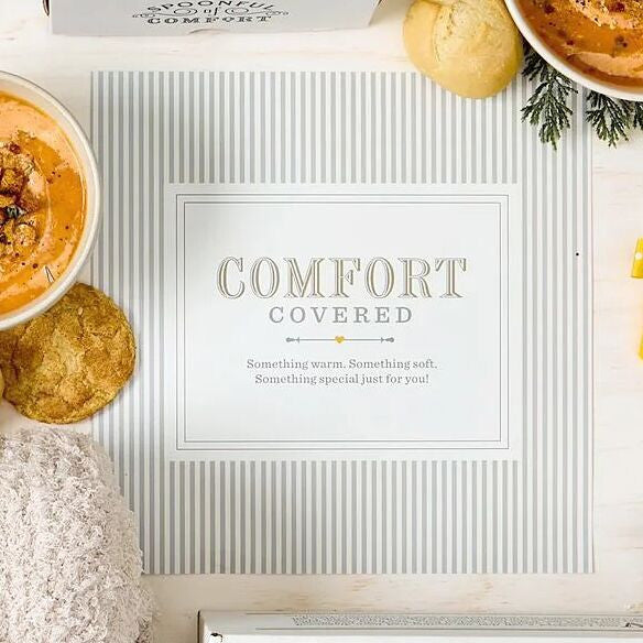Close-up of a card that reads âComfort Covered. Something warm. Something soft. Something special just for you,â surrounded by bowls of soup, rolls, cookies, and a cozy blanket.