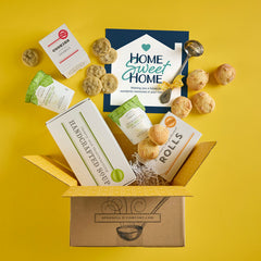 Open Spoonful of Comfort gift box with rolls, cookies, a container labeled âHandcrafted Soup,â and a card that reads âHome Sweet Home,â with rolls and cookies arranged around the box on a yellow background.