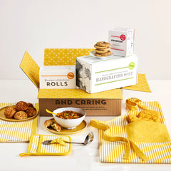 Kitchen-themed gift arrangement with a bowl of soup, rolls, cookies, boxes labeled âHandcrafted Soup,â âRolls,â and âCookies,â a metal ladle with a yellow ribbon, and yellow striped kitchen linens including an apron, towel, and oven mitts.