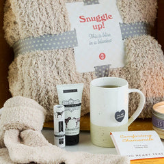 Cozy self-care gift set with a plush blanket labeled âSnuggle Up,â fuzzy socks, a mug, hand creams, a candle, and a small card arranged together on a surface.