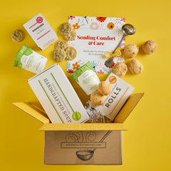 Open Spoonful of Comfort gift box with rolls, cookies, two containers of soup, a box labeled âHandcrafted Soup,â and a floral card that reads âSending Comfort & Care,â with additional rolls and cookies arranged around the box.