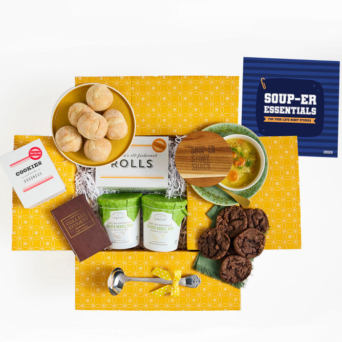 Overhead view of a yellow gift box arrangement with a bowl of soup, rolls, cookies, two containers of soup, a metal ladle, a brown book labeled âCollege Essentials,â and a blue card that reads âSoup-er Essentials.â