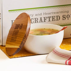 Close-up of a bowl of soup with a wooden lid resting beside it, set in front of a box labeled âHandcrafted Soup.â