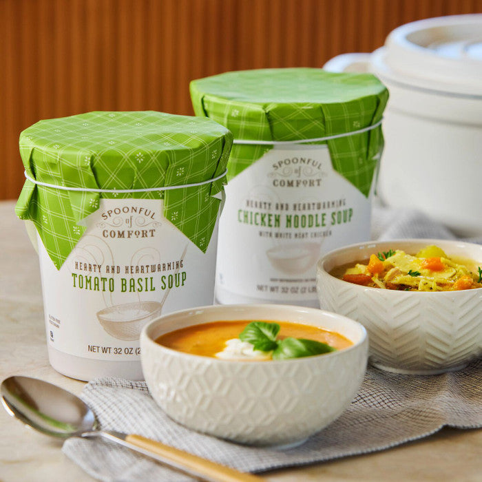 Close-up of two containers of soup with green lids labeled âChicken Noodle Soupâ and âThai Basil Soup,â with bowls of soup and a spoon arranged on a table.