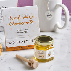 Close-up of a package labeled âComforting Chamomileâ herbal tea next to a mini jar of honey with a wooden dipper, with a mug in the background.