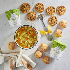 Overhead view of a pot of soup surrounded by rolls, cookies, two containers of soup with green lids, and a metal ladle with a yellow ribbon on a light surface.