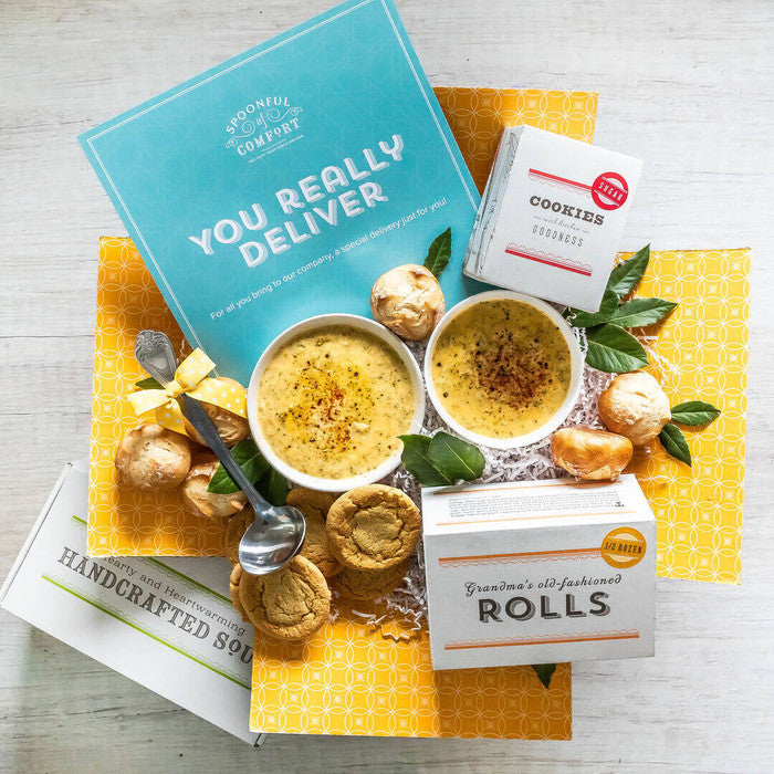 Overhead view of a yellow gift box arrangement with two bowls of soup, rolls, cookies, a container labeled âHandcrafted Soup,â a metal ladle, and a blue card that reads âYou Really Deliver.â