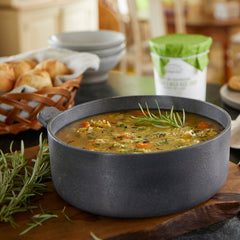 Close-up of a pot of chicken and wild rice soup garnished with herbs, set on a wooden board with rolls and bowls in the background.