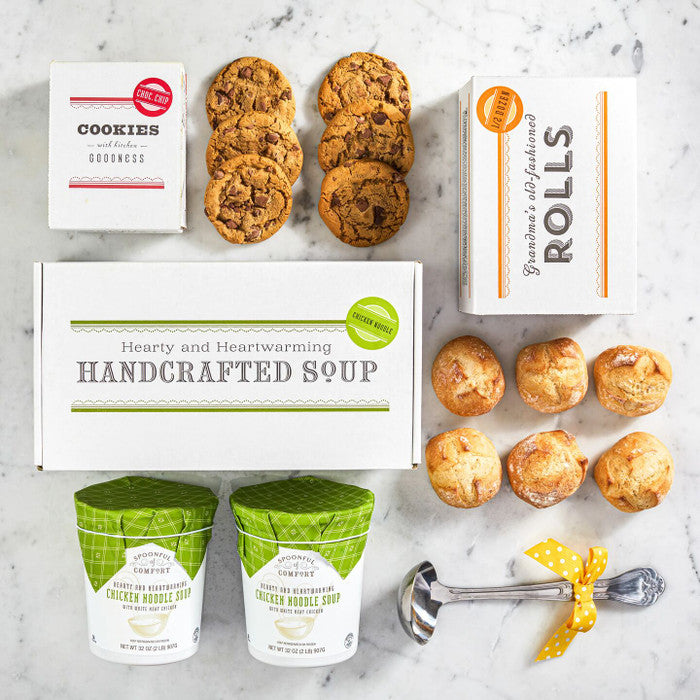 Overhead view of a soup gift set with a box labeled âHandcrafted Soup,â a box of rolls, cookies, two containers of soup, and a metal ladle with a yellow ribbon arranged on a surface.