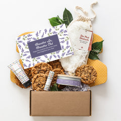 Open gift box filled with assorted cookies, a candle, lotion, lip balm, a warmer, greenery, and a floral card that reads âBreathe In, Breathe Out.â