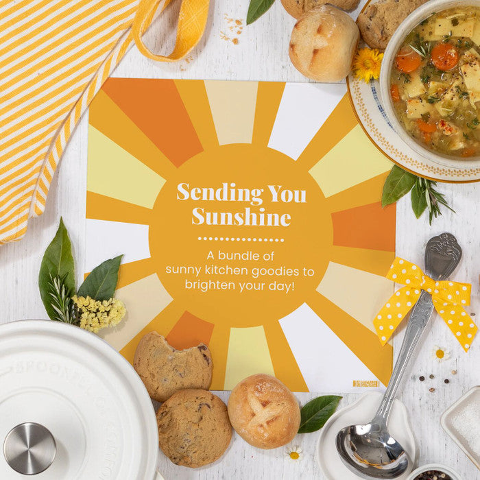 Overhead view of a sunshine-themed arrangement with soup, rolls, cookies, a metal ladle with a yellow ribbon, and a card that reads âSending You Sunshine. A bundle of sunny kitchen goodies to brighten your day.â