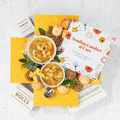 Overhead view of a yellow gift box arrangement with two bowls of soup, rolls, cookies, a container labeled âHandcrafted Soup,â a metal ladle with a yellow ribbon, and a floral card that reads âSending Comfort & Care.â
