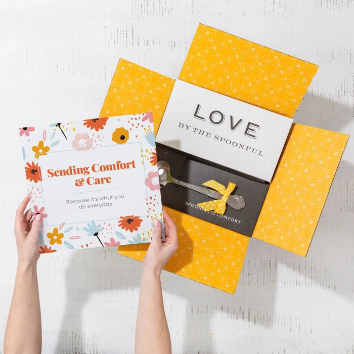 Overhead view of hands holding a floral card that reads âSending Comfort & Careâ next to an open yellow gift box with a card that reads âLove by the Spoonfulâ and a metal ladle with a yellow ribbon inside.