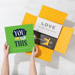 Overhead view of hands holding a green card that reads âYouâve Got Thisâ next to an open yellow gift box with a card that reads âLove by the Spoonfulâ and a metal ladle with a yellow ribbon inside.