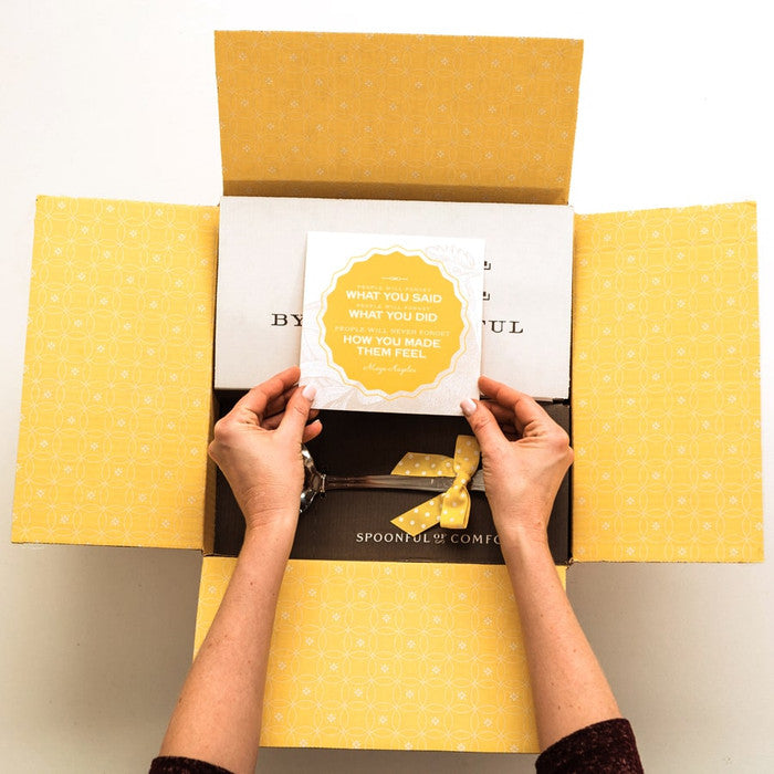 A Spoonful of Comfort shipping box opened to display a yellow printed interior. Hands reaching into a box holding an insert card that reads " people will forget what you said, people will forget what you did, but people will never forget how you made them feel.â
â Maya Angelou"