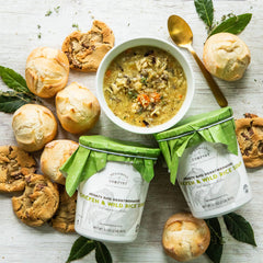 A flat lay display of Chicken & Wild Rice soup in a bowl and two Spoonful of Comfort soup containers, rolls and cookies.