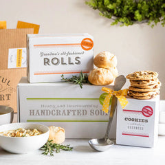 Soup gift set displayed with a bowl of soup, rolls, cookies, boxes labeled âChicken Soup,â âRolls,â and âCookies,â and a metal ladle with a yellow ribbon.