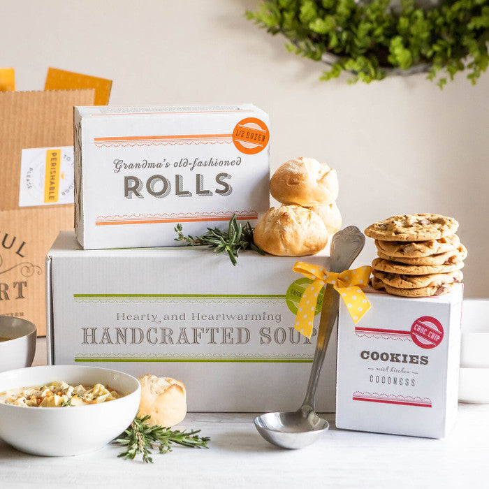 Soup gift set displayed with a bowl of soup, rolls, cookies, boxes labeled âHandcrafted Soup,â âRolls,â and âCookies,â and a metal ladle with a yellow ribbon.