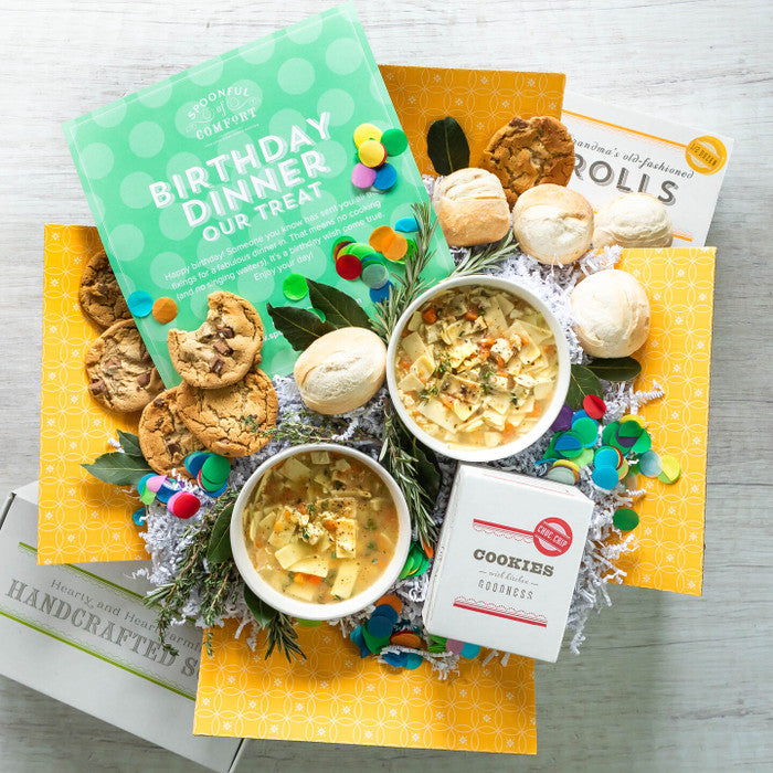Overhead view of a birthday-themed gift arrangement with two bowls of soup, rolls, cookies, a container labeled âHandcrafted Soup,â a metal ladle, colorful confetti, and a green card that reads âBirthday Dinner Our Treat.â
