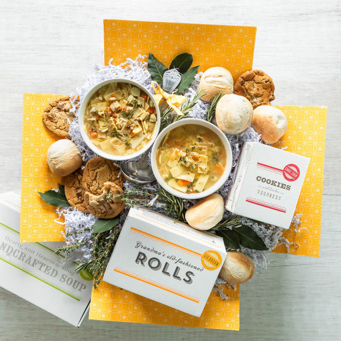 Overhead view of a yellow gift box arrangement with a bowl of soup, rolls, cookies, a box of cookies, a box of rolls, and a container labeled âHandcrafted Soup,â arranged with greenery.