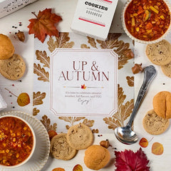 Overhead view of a fall-themed gift arrangement with two bowls of soup, rolls, a container labeled âHandcrafted Soup,â a metal ladle with a yellow ribbon, autumn leaves, and a card that reads âUp and Autumn.â