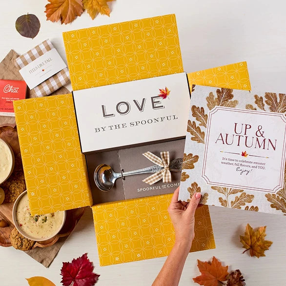 Overhead view of a fall-themed gift arrangement with two bowls of soup, rolls, a container labeled âHandcrafted Soup,â a metal ladle with a yellow ribbon, autumn leaves, and a card that reads âUp and Autumn.â