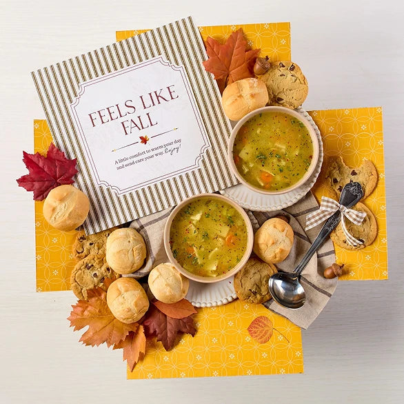 Overhead view of a fall-themed arrangement with two bowls of soup, rolls, cookies, a box labeled âHandcrafted Soup,â a box of rolls, a metal ladle with a yellow ribbon, autumn leaves, and a card that reads âFeels Like Fall.â