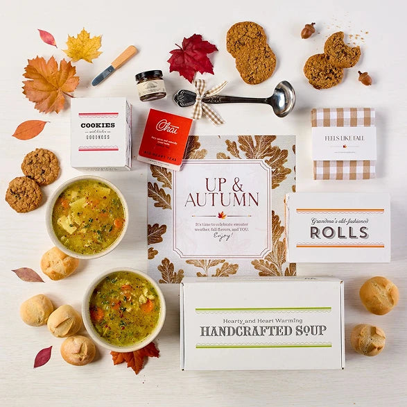 Overhead view of a fall-themed gift arrangement with two bowls of soup, rolls, a container labeled âHandcrafted Soup,â a metal ladle with a yellow ribbon, autumn leaves, and a card that reads âUp and Autumn.â