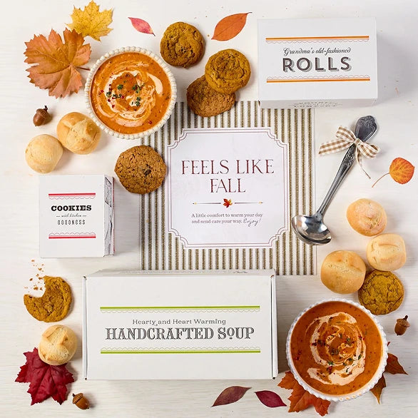 Overhead view of a fall-themed arrangement with two bowls of soup, rolls, cookies, a box labeled âHandcrafted Soup,â a box of rolls, a metal ladle with a yellow ribbon, autumn leaves, and a card that reads âFeels Like Fall.â