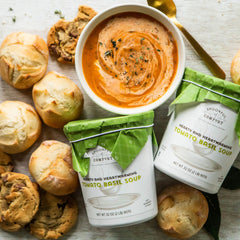 Close-up of a bowl of tomato soup with rolls and cookies, alongside two containers labeled âTomato Basil Soupâ with green lids.
