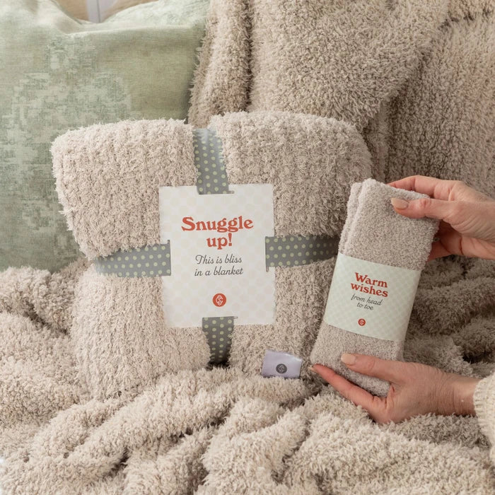 Close-up of a cozy plush blanket labeled âSnuggle Upâ and a pair of soft socks labeled âWarm Wishes,â held by hands against a textured blanket background.