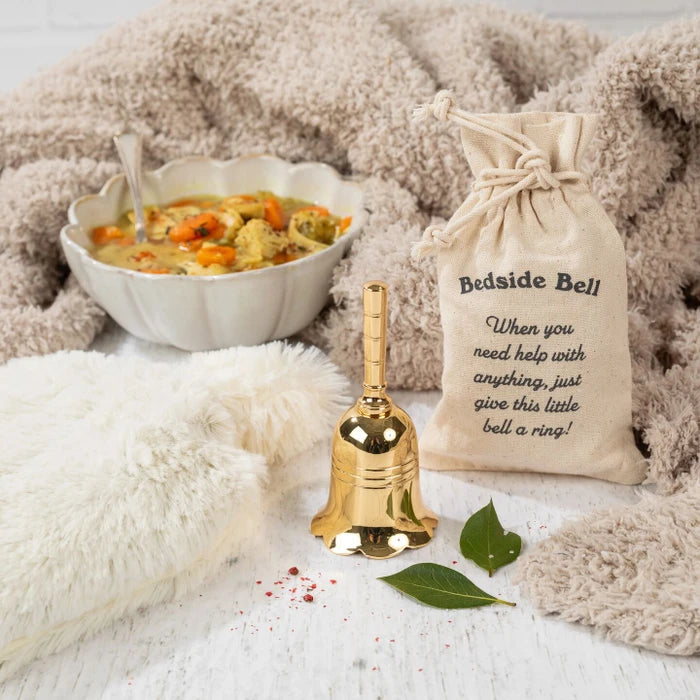 Close-up of a gold bedside bell next to a drawstring bag labeled âBedside Bell,â with a bowl of soup and a cozy blanket in the background.