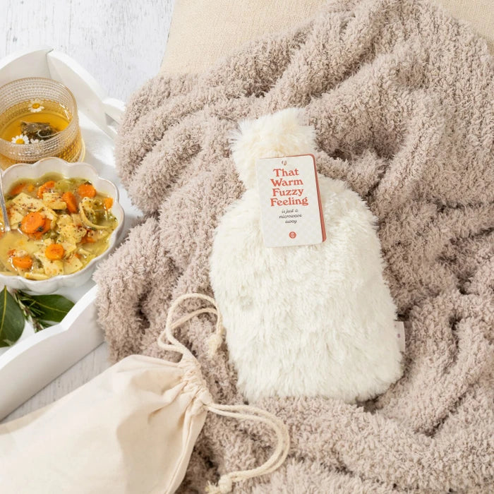 Close-up of a cozy plush blanket with a âWarm Fuzziesâ warm/cool pack on top, next to a tray with a bowl of soup and a glass of tea.
