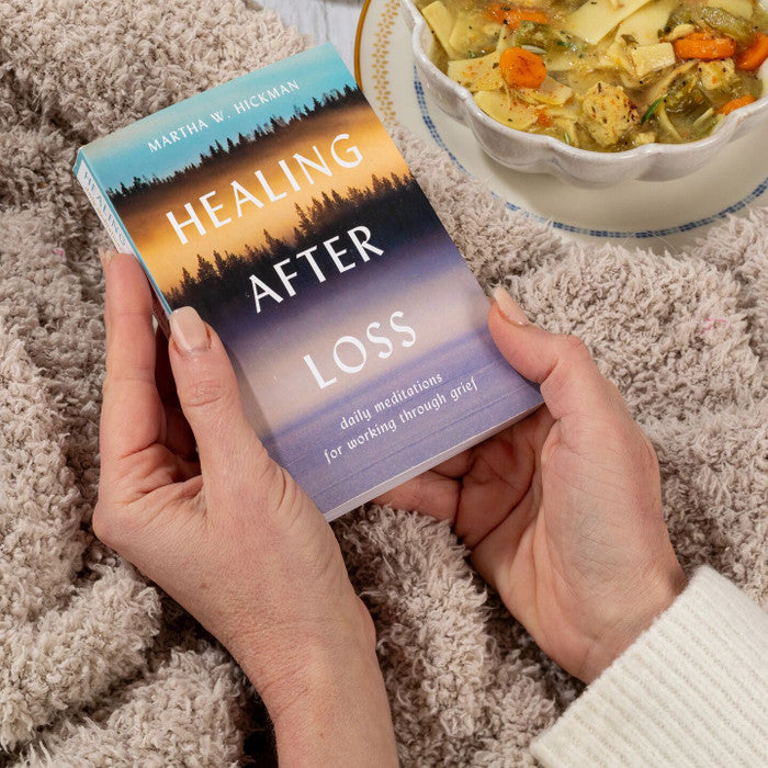 Hands holding a book titled âHealing After Loss,â resting on a cozy blanket with a bowl of soup nearby.