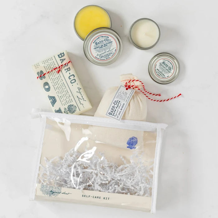 Close-up of a clear toiletry bag filled with self-care items including a soothing bath soak, triple-milled bar soap, moisturizing hand salve, and a scented candle by Barr-Co., arranged on a light surface.