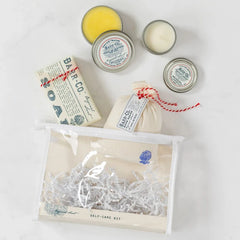 Close-up of a clear toiletry bag filled with self-care items including a soothing bath soak, triple-milled bar soap, moisturizing hand salve, and a scented candle by Barr-Co., arranged on a light surface.