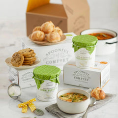 A sympathy soup gift package with soup containers, a bowl of soup, cookies, dinner rolls, and a Spoonful of Comfort box arranged on a light kitchen surface.