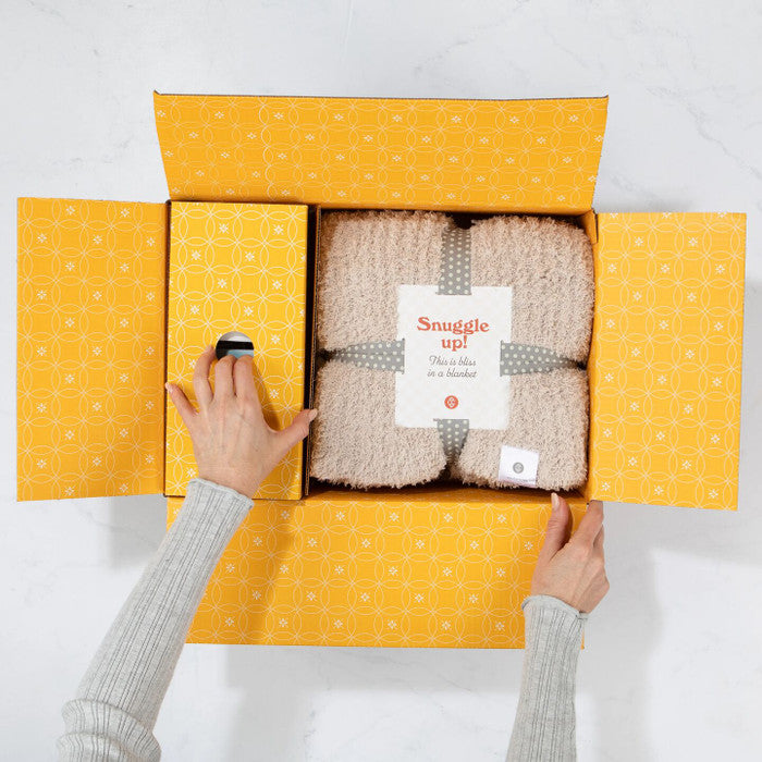 Overhead view of hands opening a yellow gift box revealing a cozy knit blanket labeled âSnuggle Upâ inside.
