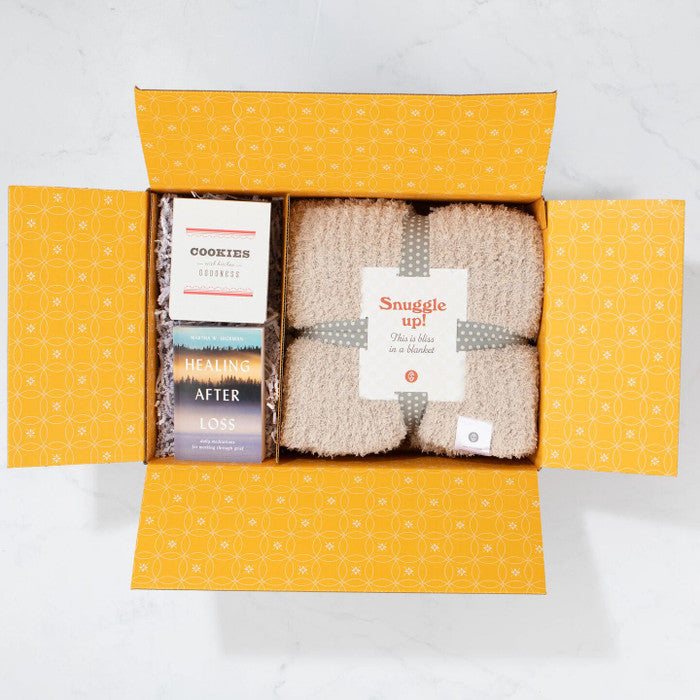 Open yellow gift box displaying a cozy knit blanket labeled âSnuggle Up,â alongside a box of cookies and a book titled âHealing After Loss.â