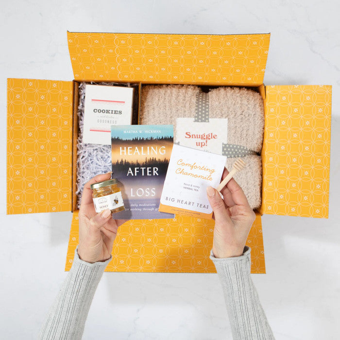 Overhead view of hands holding a package of chamomile tea and a book titled âHealing After Lossâ above an open yellow gift box containing a cozy knit blanket labeled âSnuggle Upâ and a box of cookies.