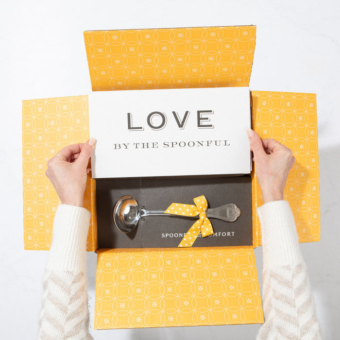 Open yellow gift box displaying a card that reads âLove by the Spoonfulâ and a metal ladle with a yellow ribbon inside.