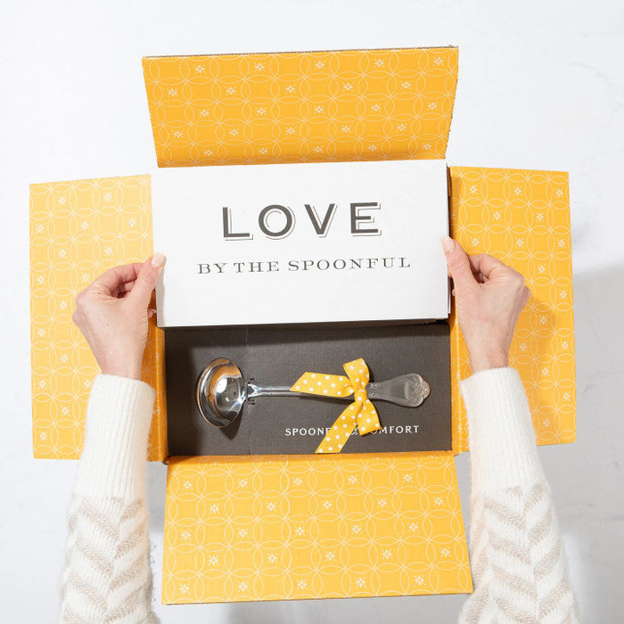 Open yellow gift box displaying a card that reads âLove by the Spoonfulâ and a metal ladle with a yellow ribbon inside.