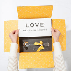 Open yellow gift box displaying a card that reads âLove by the Spoonfulâ and a metal ladle with a yellow ribbon inside.