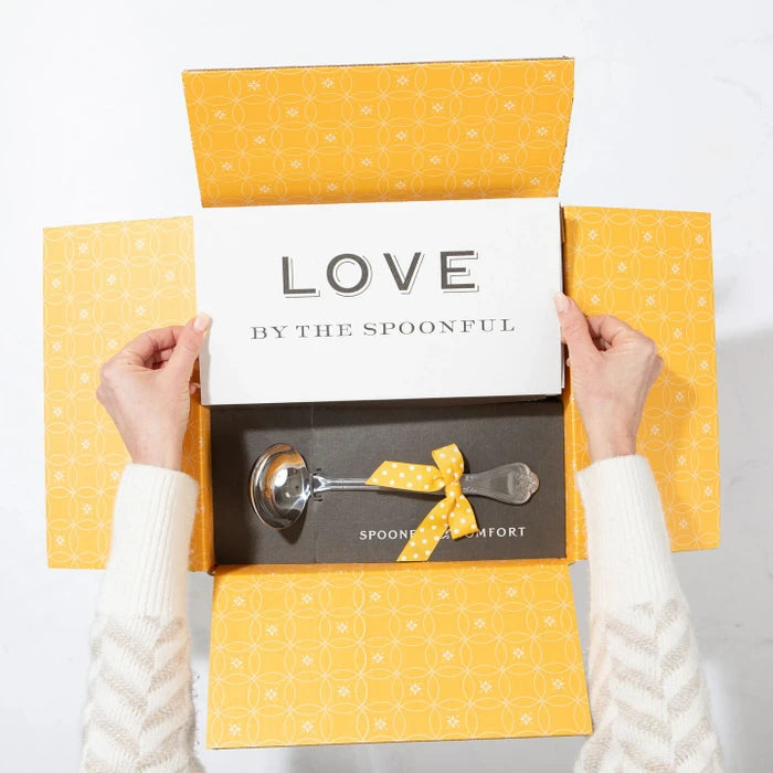 Open yellow gift box displaying a card that reads âLove by the Spoonfulâ and a metal ladle with a yellow ribbon inside.