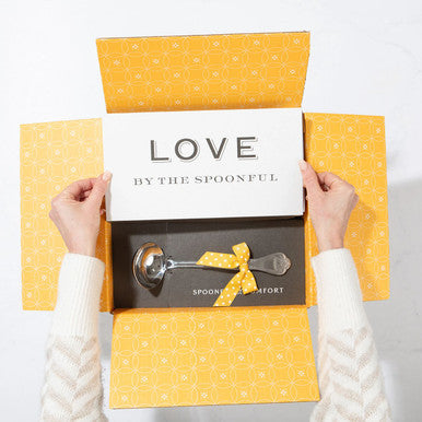 Open yellow gift box displaying a card that reads âLove by the Spoonfulâ and a metal ladle with a yellow ribbon inside.